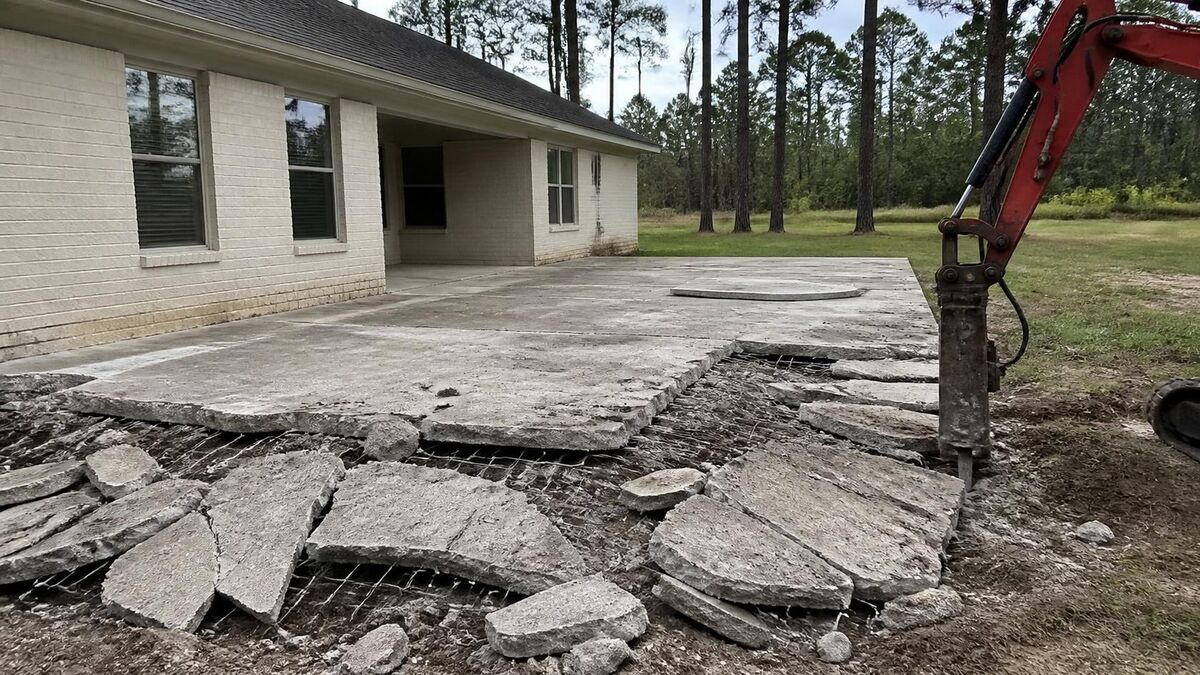 A mini excavator hammer breaking up an old concrete patio slab behind a ranch house