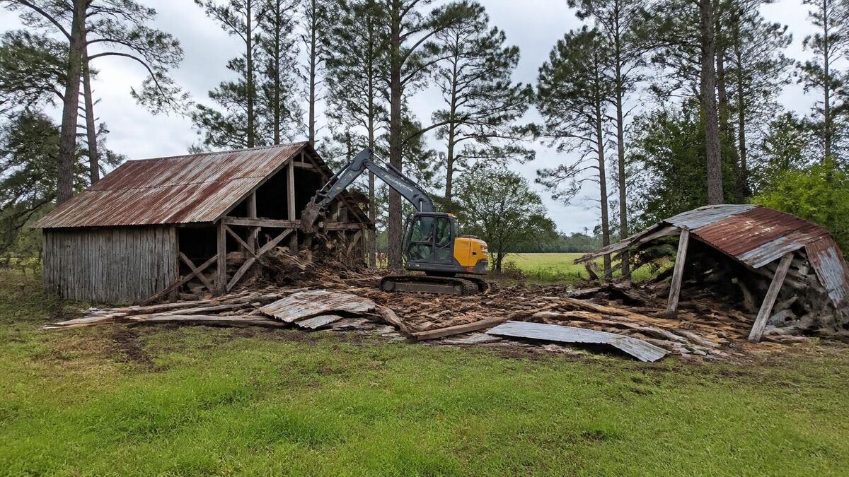 A compact excavator pulling down an old metal-roofed barn and shed on rural acreage